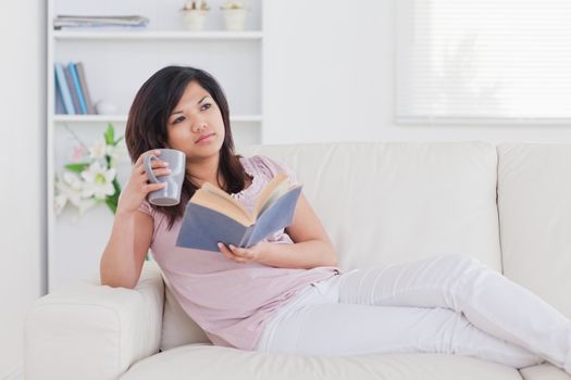 Woman lying on a couch while holding a book and a mug in a living room
