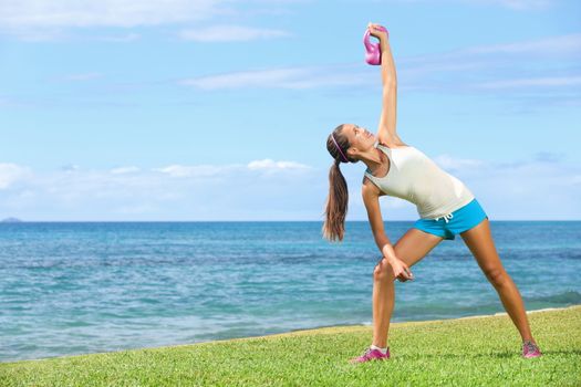 Crossfit exercise woman using kettlebell in fitness strength training workout outside on grass by the ocean. Beautiful young fit instructor and fitness model.