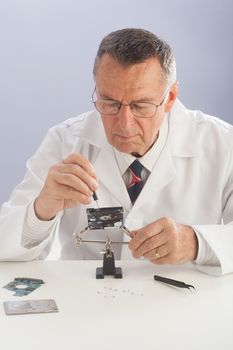 An older male wearing a white lab coat and reaparing electronic equipments, like a technician or a repair man.