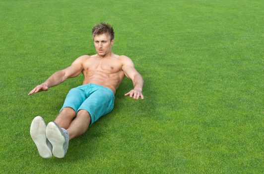 Young man training outdoors and doing sit-ups on green sports field.