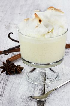 Rice pudding in a glass bowl topped with meringue. Star anise, cinnamon bark and vanilla pods are in the background.