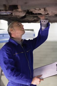 Mechanic touching the below of a car while holding a clipboard in a garage