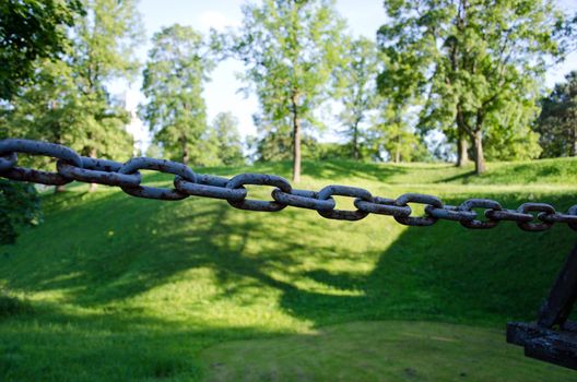 Closeup of rusty steel chain and green mound with trees on blurred background.