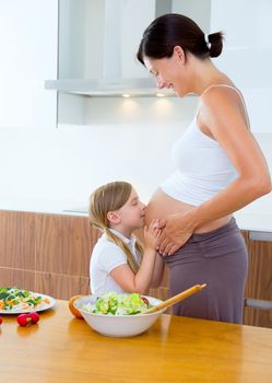 Beautiful pregnant mother with her daughter at kitchen kissing baby in belly