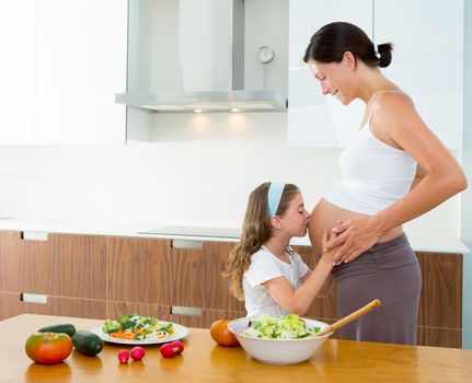 Beautiful pregnant mother with her daughter at kitchen kissing baby in belly