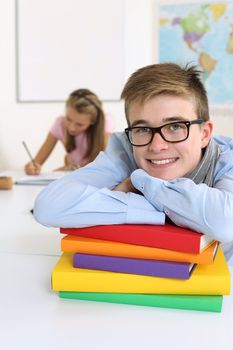 Photo of a handsome young male smiling in his classroom with a female in the background writing and reading.