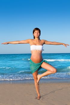Beautiful pregnant woman practicing yoga in blue beach sand on summer vacation