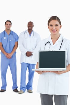 Smiling doctor showing laptop with colleagues behind her against a white background