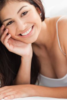 A close up shot of a smiling woman who is looking forward with a hand resting on her cheek and her other hand upon a pillow.