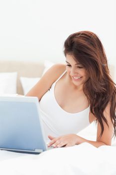 A close up shot of a woman lying on the bed as she smiles while she uses her laptop. 