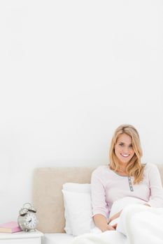 A Vertical shot of a smiling woman sitting in bed against the headboard with the alarm clock on a desk beside her.