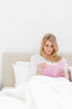 A closer shot of a woman in bed sitting upright as she reads her book.