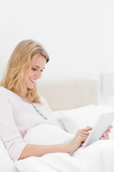 A side view shot of a woman sitting in bed with a tablet pc that she is watching while smiling.