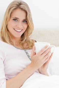 A close up shot of a woman holding a cup in her hands near to her head as she looks forward with a smile.