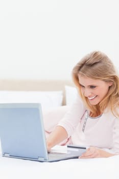 A close up shot of a smiling woman who is looking at her laptop as she lies upon her bed.