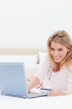 A smiling woman lying on her bed as she uses her laptop while looking straight ahead.