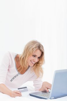 A woman lying on her bed is smiling as she is ordering items online with her credit card and laptop