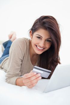 A close up shot of a smiling woman lying on the bed with a credit card and tablet in her hands.