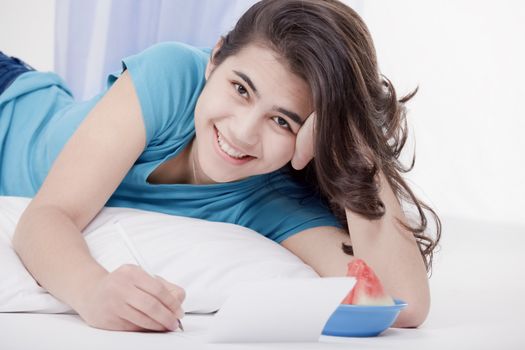 Relaxed and happy biracial teen girl or young woman lying on floor pillow writing a letter or note.