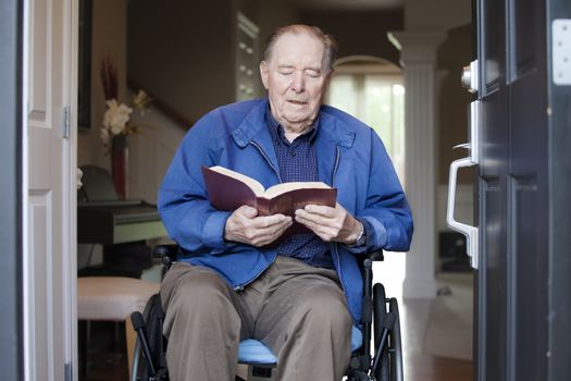 Elderly 90 yr old man in wheelchair at his front door, reading a Bible
