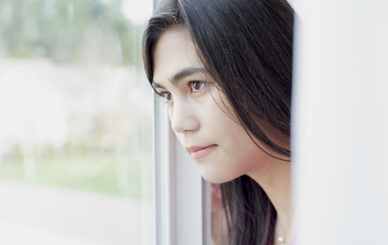 Side profile of teen girl or young woman looking out sunny window, sad or lonely expression