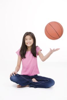 Ten year old Asian girl sitting on floor throwing basketball up in the air, isolated on white