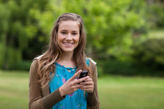 Smiling teenage standing in a park while sending a text with her cellphone