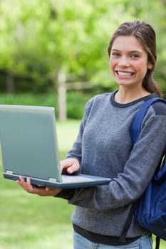 Happy teenage holding her laptop while looking at the camera and standing in a park