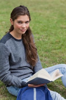 Young calm girl holding a book while sitting on the grass next to her backpack
