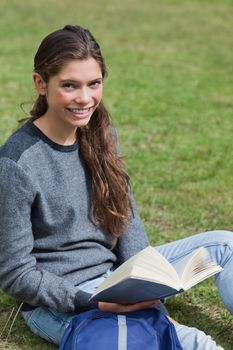 Smiling young girl holding an open book while sitting down on the grass in a park