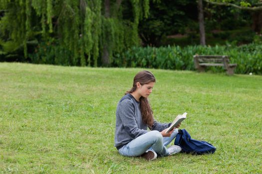 Young girl sitting cross-legged on the grass in a park while reading a book