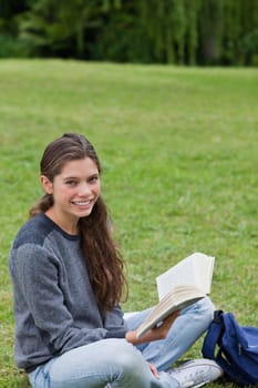Young smiling girl sitting cross-legged on the grass while holding a book in a park