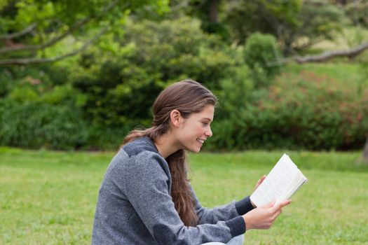 Side view of a young girl showing a great smile while reading a book on the grass