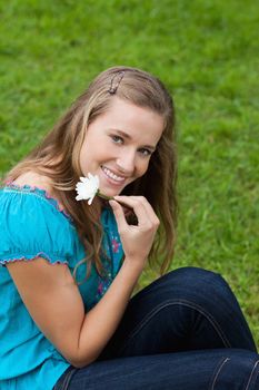 Smiling young girl holding a flower next to her face while sitting in a park