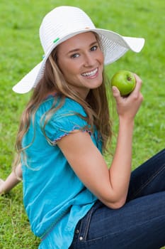 Attractive young girl sitting on the grass while eating a green apple
