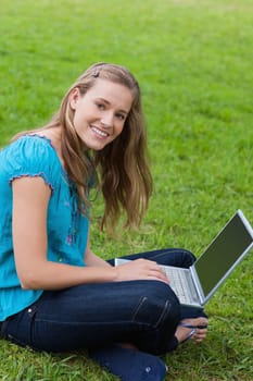 Young smiling girl sitting in a park while looking at the camera and using her laptop 