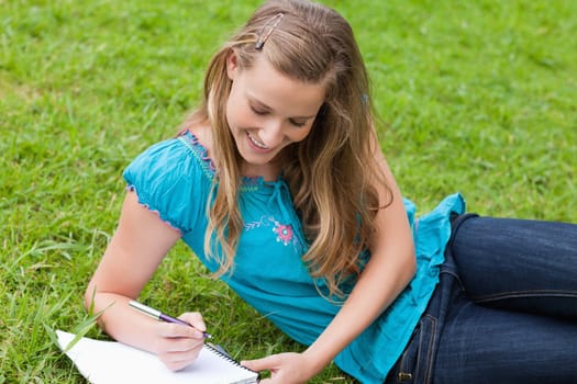 Young girl lying on the grass in a park while writing on a notebook and smiling