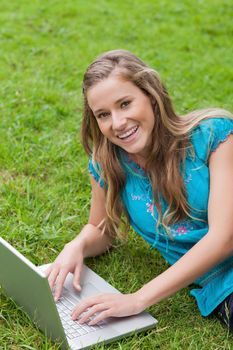 Smiling student lying on the side on the grass while using her laptop