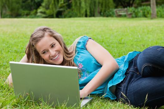 Young student lying on the side in a park while working on her laptop and smiling