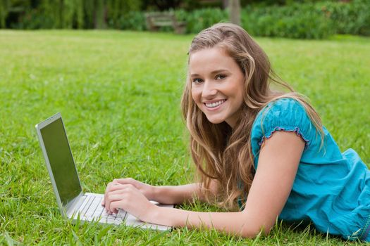 Happy teenager using her laptop while lying in a park and looking at the camera