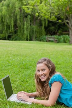 Young girl lying on the grass in a park while typing on her laptop and laughing