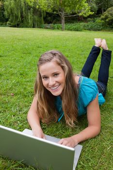 Happy student looking at the camera while lying in a park while using her laptop