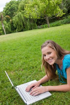 Smiling young girl lying on the grass in a public garden while working on her laptop