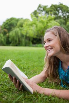Young smiling woman lying in a parkland while holding a book and looking away