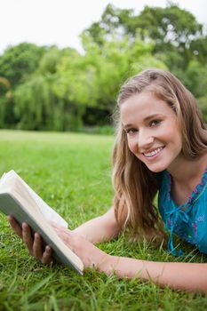 Smiling teenager holding a book while lying in a parkland and looking at the camera