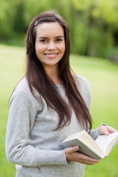 Happy girl standing upright in a park while looking at the camera and holding a book