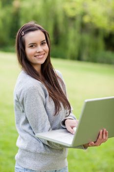 Young happy woman holding her laptop while standing in a parkland and smiling
