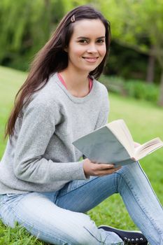 Young smiling girl holding a book while sitting on the grass and looking at the camera