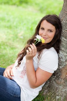 Young woman smelling flowers while sitting against a tree and looking at the camera