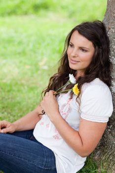 Young relaxed woman leaning against a tree while holding two beautiful flowers